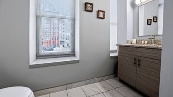 A bathroom with a toilet, a window with blinds, a vanity with a mirror, and a tiled floor.