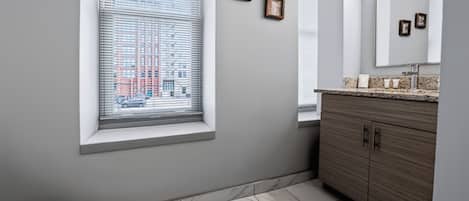 A bathroom with a toilet, a window with blinds, a vanity with a mirror, and a tiled floor.