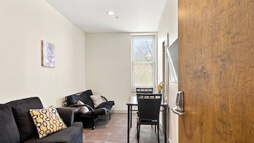 A room with a black sofa, a dining table with chairs, a wooden door, and a window with a view of trees.