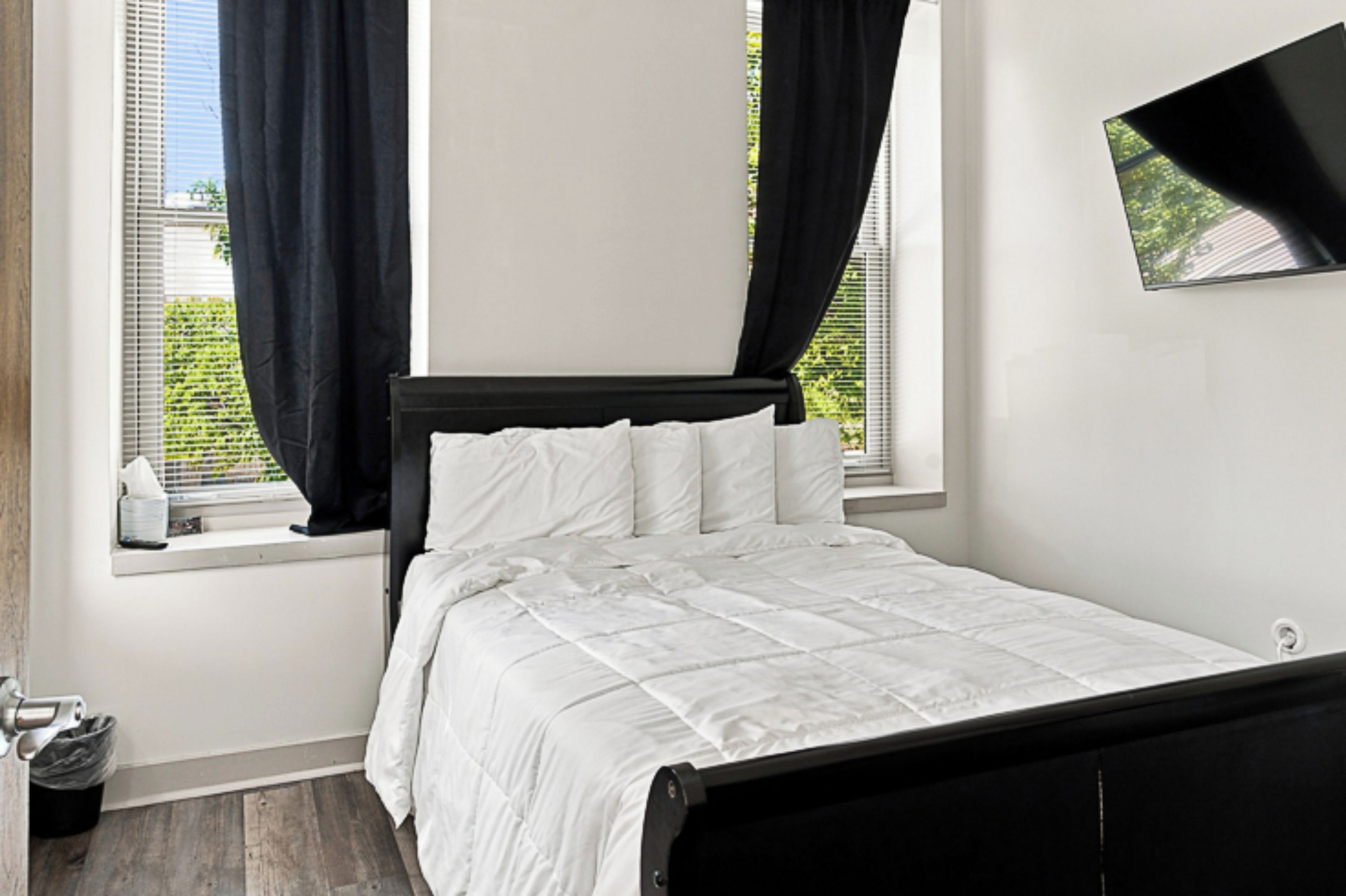 A bedroom with a bed, a window with curtains, a TV mounted on the wall, and a small trash bin.
