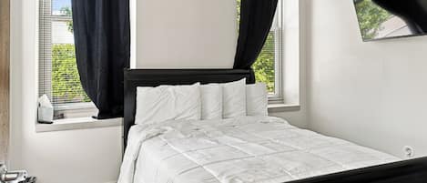 Ein Schlafzimmer mit Bett, einem Fenster mit Vorhängen, einem an der Wand befestigten Fernseher und einer kleinen Mülltonne.