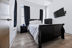 A bedroom with a bed, a nightstand, a dresser, a TV mounted on the wall, and a window with curtains.