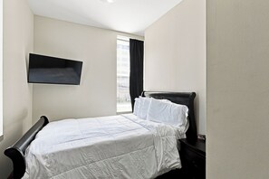 A bedroom with a bed, a nightstand, a wall-mounted shelf, and a window with curtains.