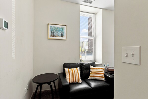 A living room with a black leather sofa, a small round table, a framed painting on the wall, and a window with blinds.