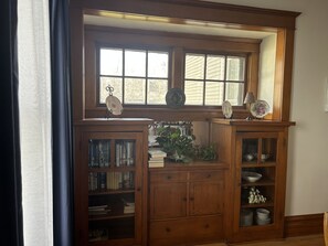 A wooden cabinet with glass doors, books, and decorative plates. - Ada’s Guesthouse- historic home close to lake, shopping, parks, trails, BVU (Storm Lake)