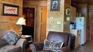 L’intérieur chaleureux d’un chalet présente un plafond en bois, un ventilateur de plafond, un fauteuil en cuir marron, une table basse en bois avec des magazines, un téléviseur et un coin cuisine équipé d’un four à micro-ondes et d’un réfrigérateur.