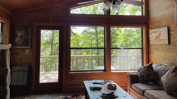 L’intérieur d’un chalet en bois, avec un ventilateur de plafond, de grandes fenêtres, un canapé et une table sur laquelle sont posés des livres.