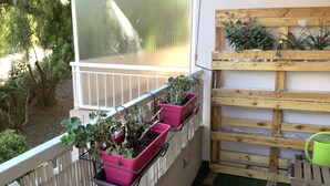 Un balcón con plantas en macetas, un banco de madera y un cubo verde.