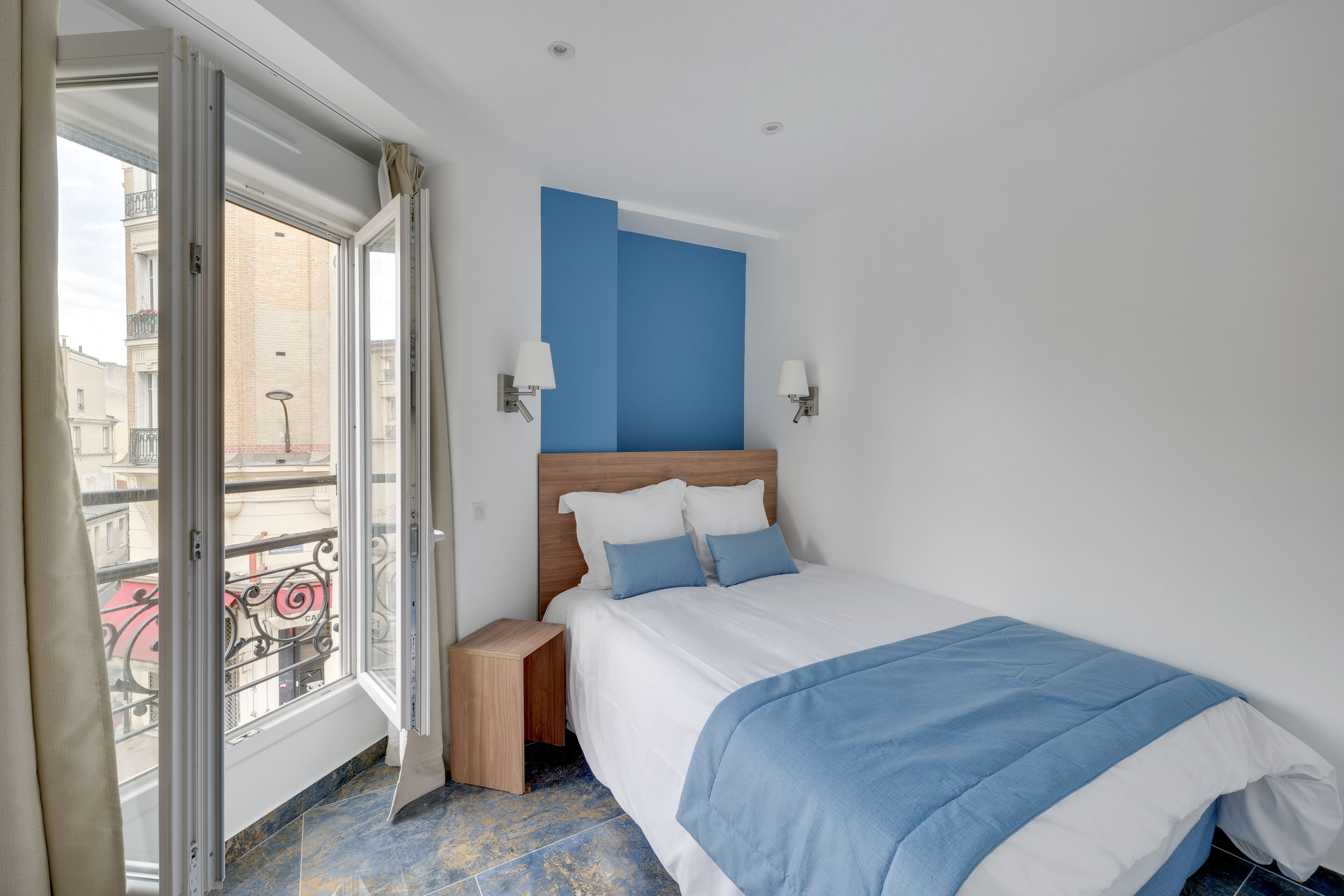 A bedroom with a bed, a nightstand, a window with a view of buildings, and a blue accent wall.