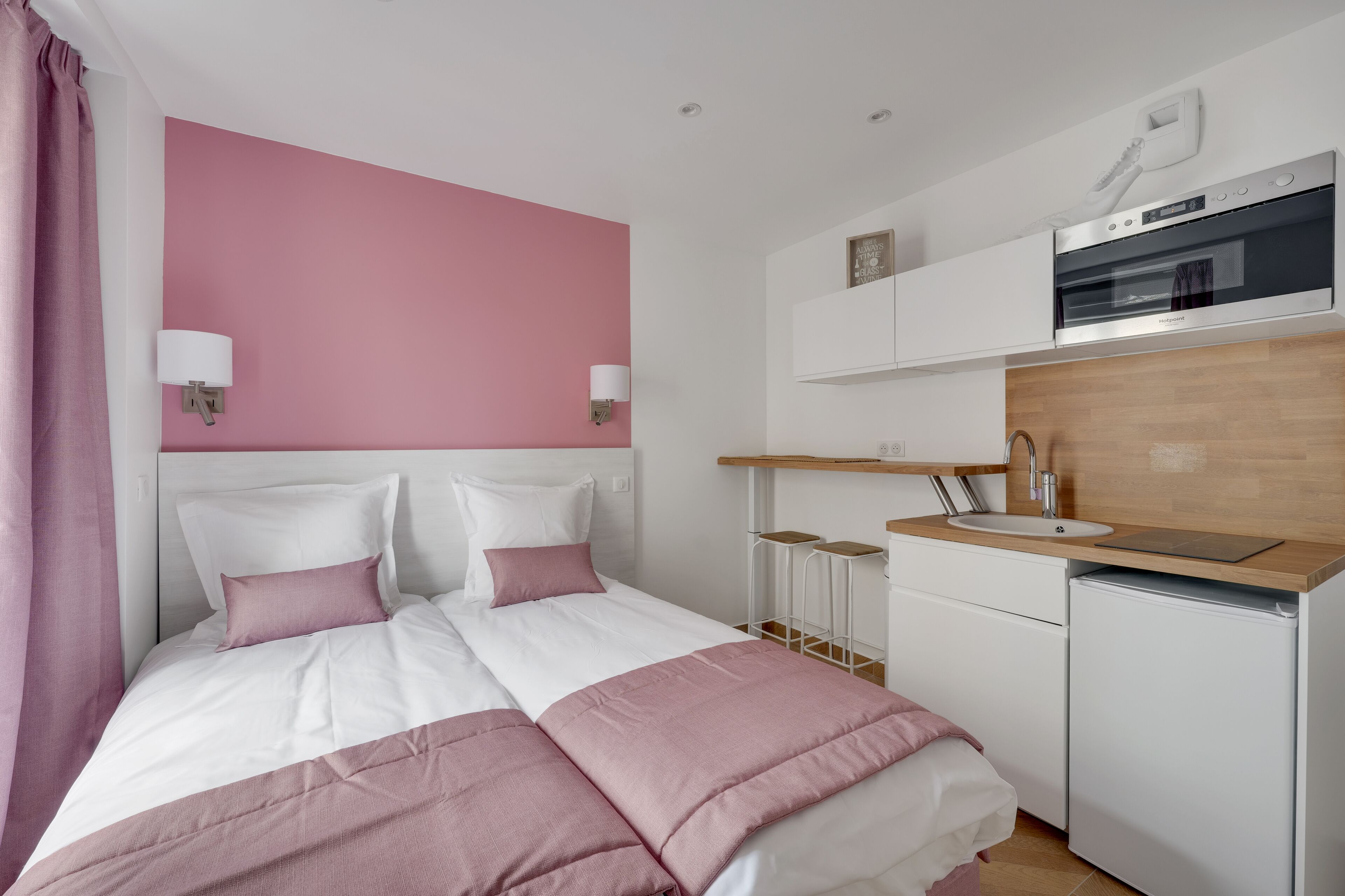 A bedroom with a bed, bedside tables, a built-in desk, a mini-fridge, and a kitchenette with a microwave and oven.