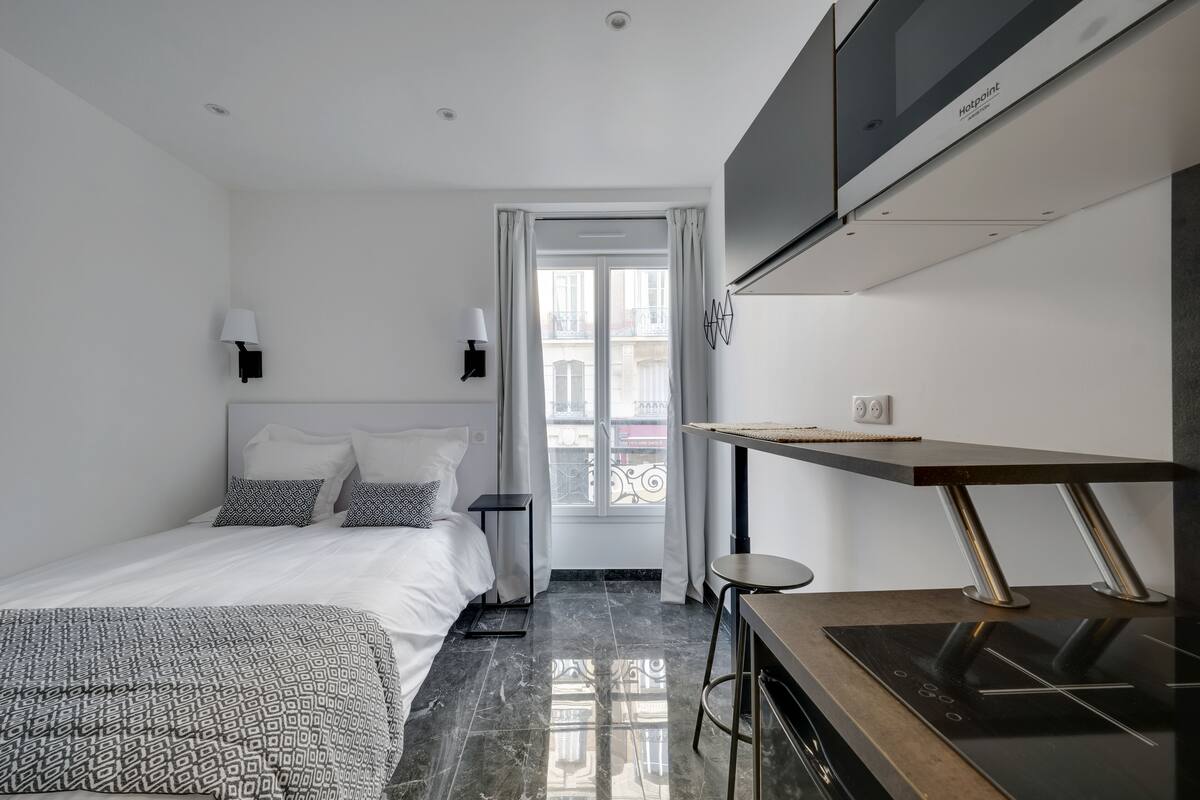 Ein modernes Hotelzimmer mit einem Bett, einem Schreibtisch mit integriertem Kochfeld, einem an der Wand montierten Fernseher und einem Fenster mit Vorhängen.
