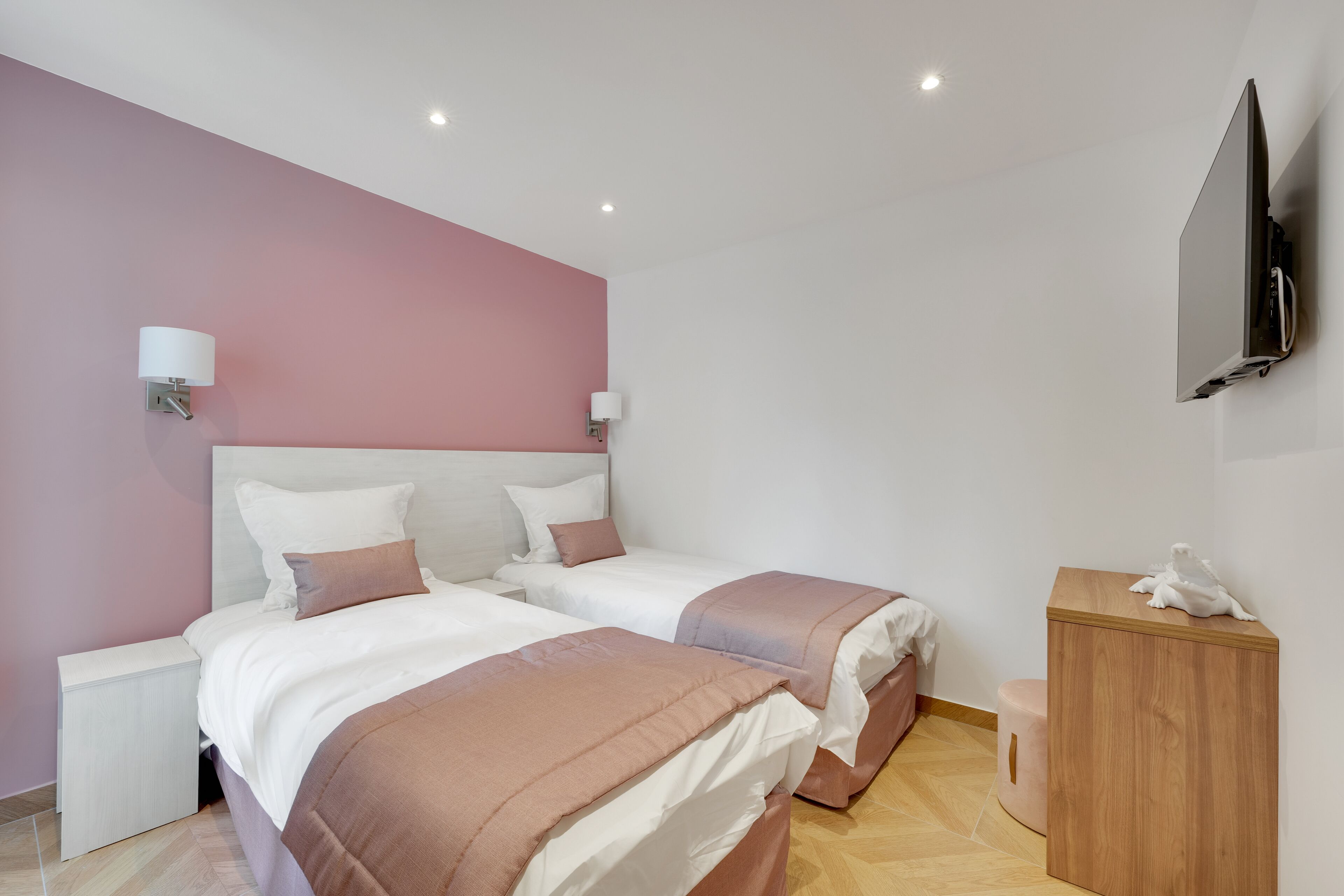 A modern hotel room with two beds, a wooden nightstand, a small wooden cabinet, and a flat-screen TV mounted on the wall.