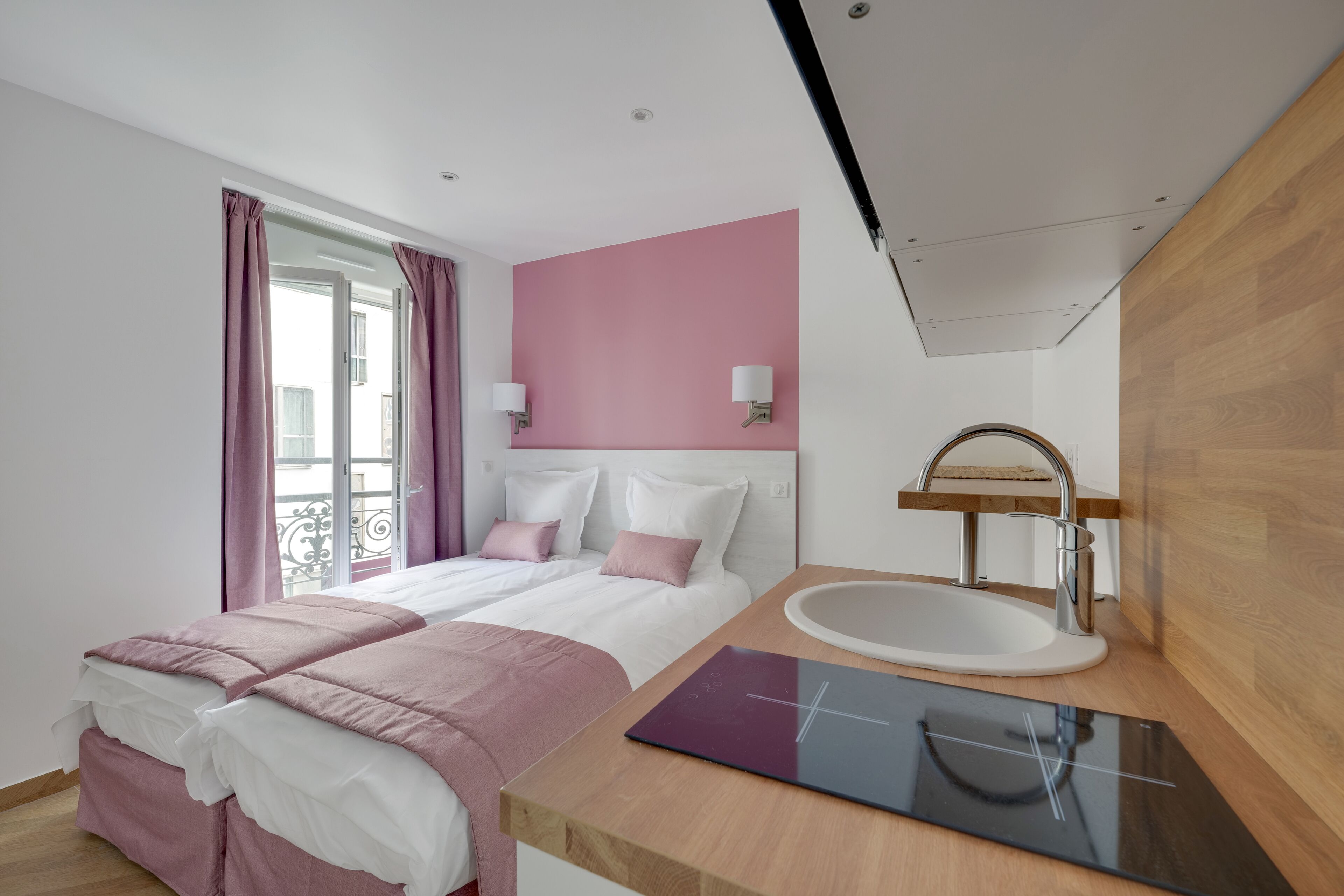 A modern hotel room with a bed, a bedside table, a sink, and a window with curtains.