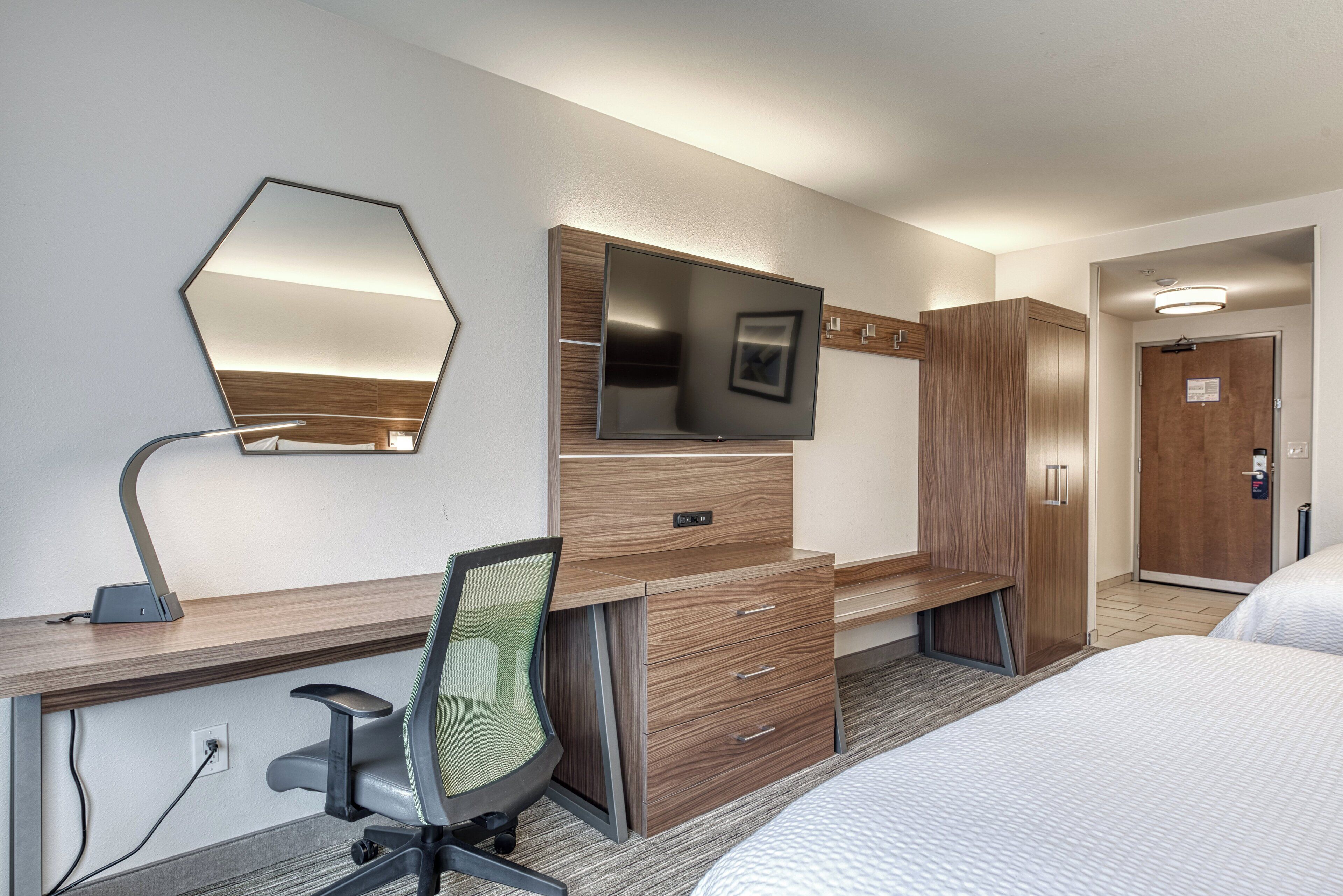 A hotel room with a bed, a desk with a chair, a television, a wardrobe, and a mirror.