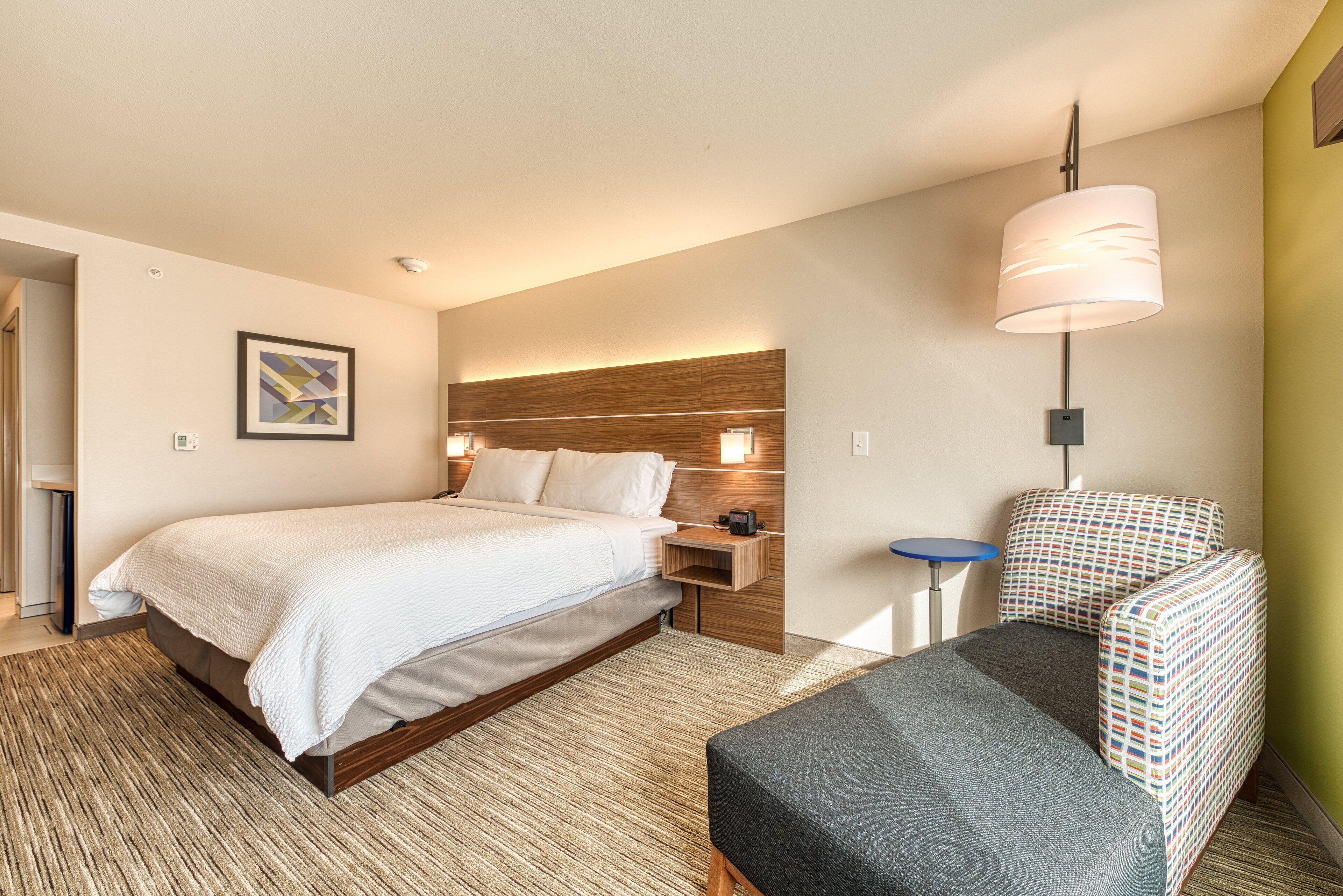 Ein modernes Hotelzimmer mit Bett, Nachttisch, Lampe, Stuhl und einem an der Wand befestigten Kunstwerk.