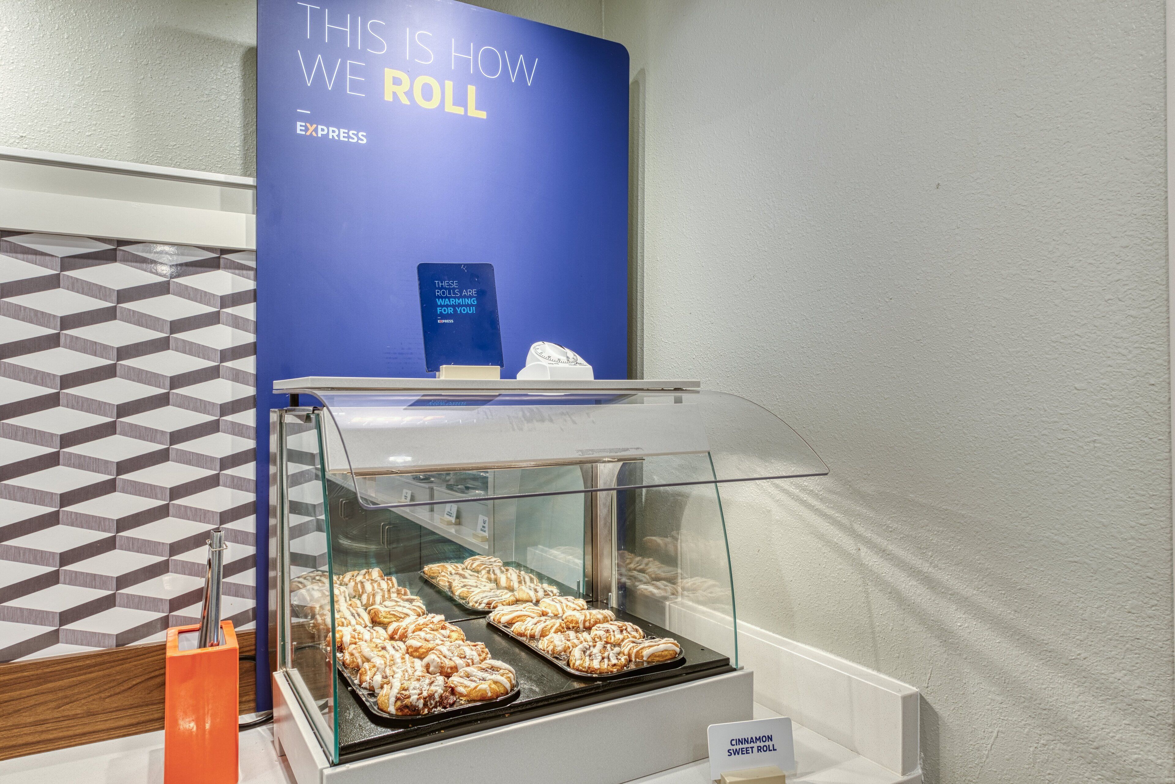 A bakery display with cinnamon sweet rolls, a sign for Express, and a geometric patterned wall.