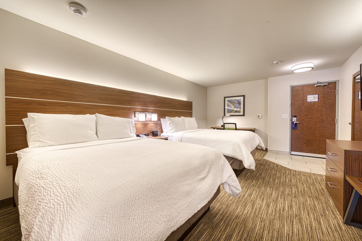 A hotel room with two beds, a wooden headboard, a desk, a chair, a nightstand, a lamp, and a door.
