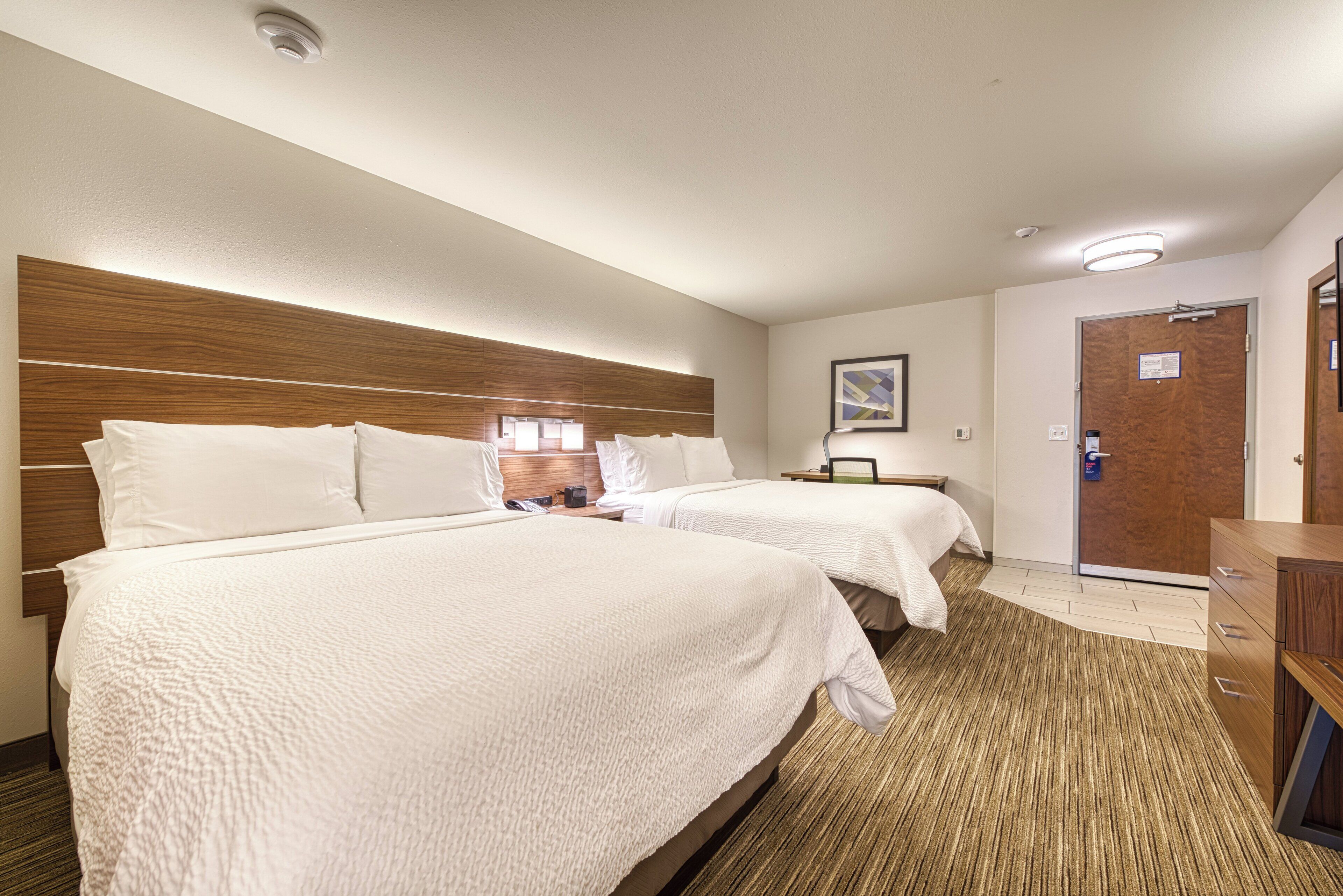 Habitación de hotel con dos camas, cabecera de madera, mesita de noche con lámpara, una puerta y un pequeño escritorio con silla.