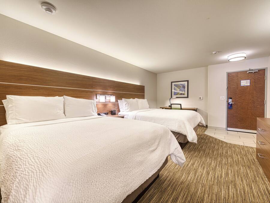 Habitación de hotel con dos camas, cabecera de madera, mesita de noche con lámpara, una puerta y un pequeño escritorio con silla.