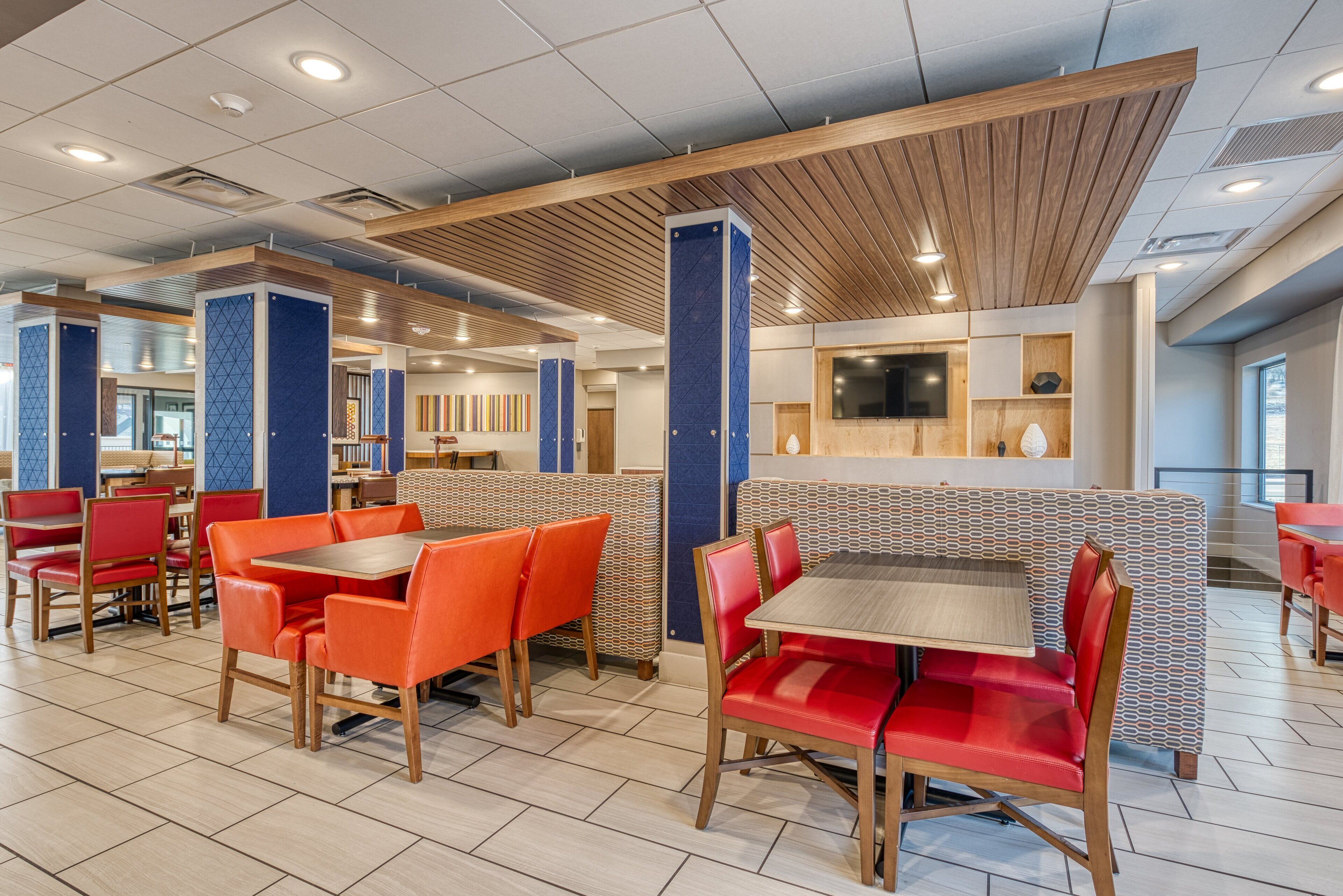 A modern dining area with booths and tables, wooden ceiling, and a TV screen on the wall.