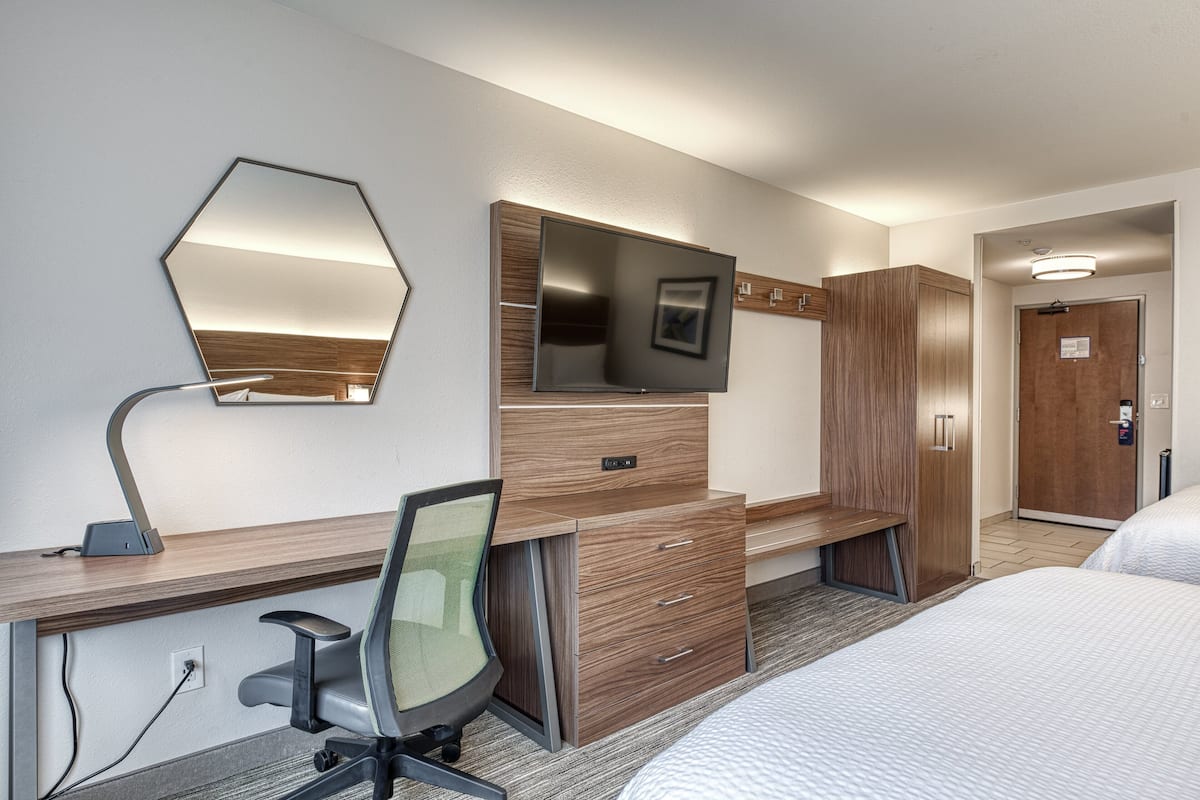 A hotel room with a wooden desk, a flat-screen TV, a mirror, a bed, and a wardrobe.