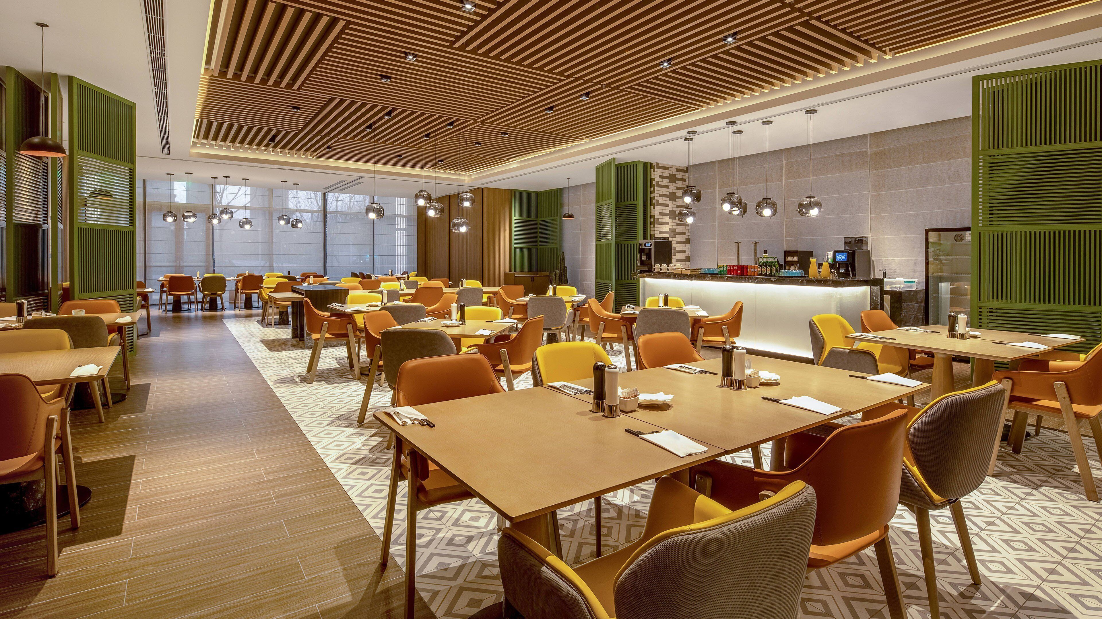 A modern restaurant with wooden ceiling panels, yellow and orange seating, and a central bar area.