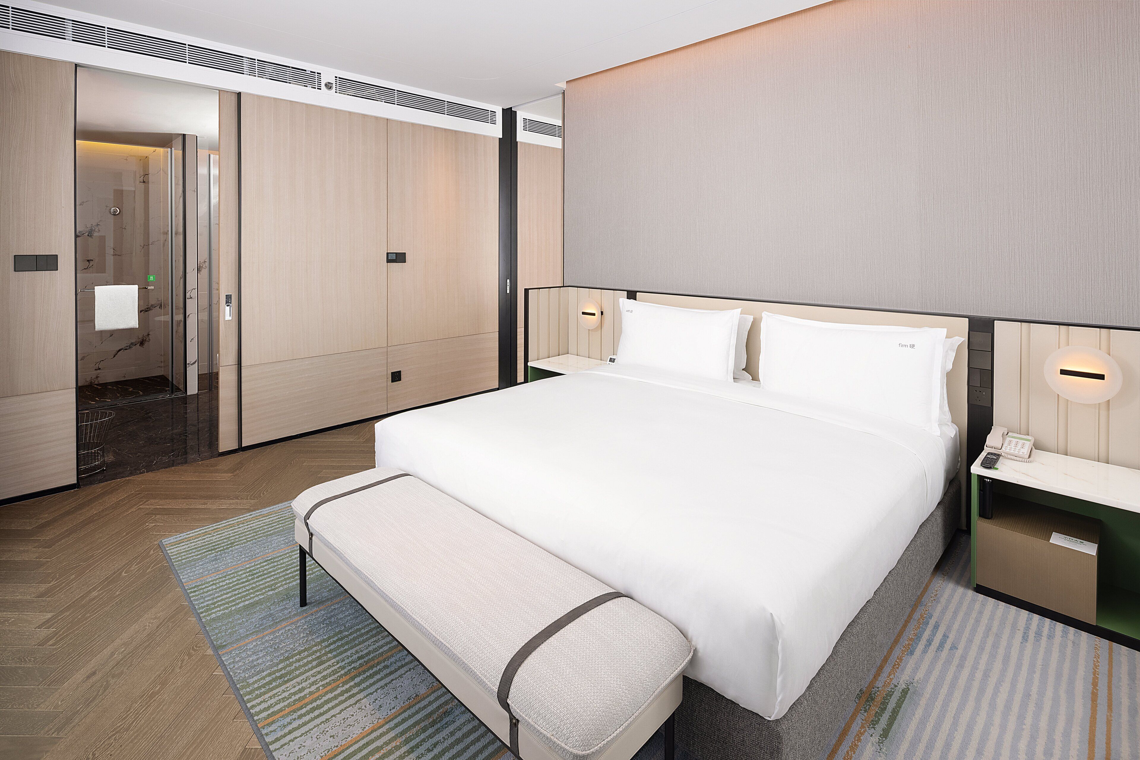 A modern hotel room with a large bed, a bench, a bedside table with a phone, and a wall-mounted light.
