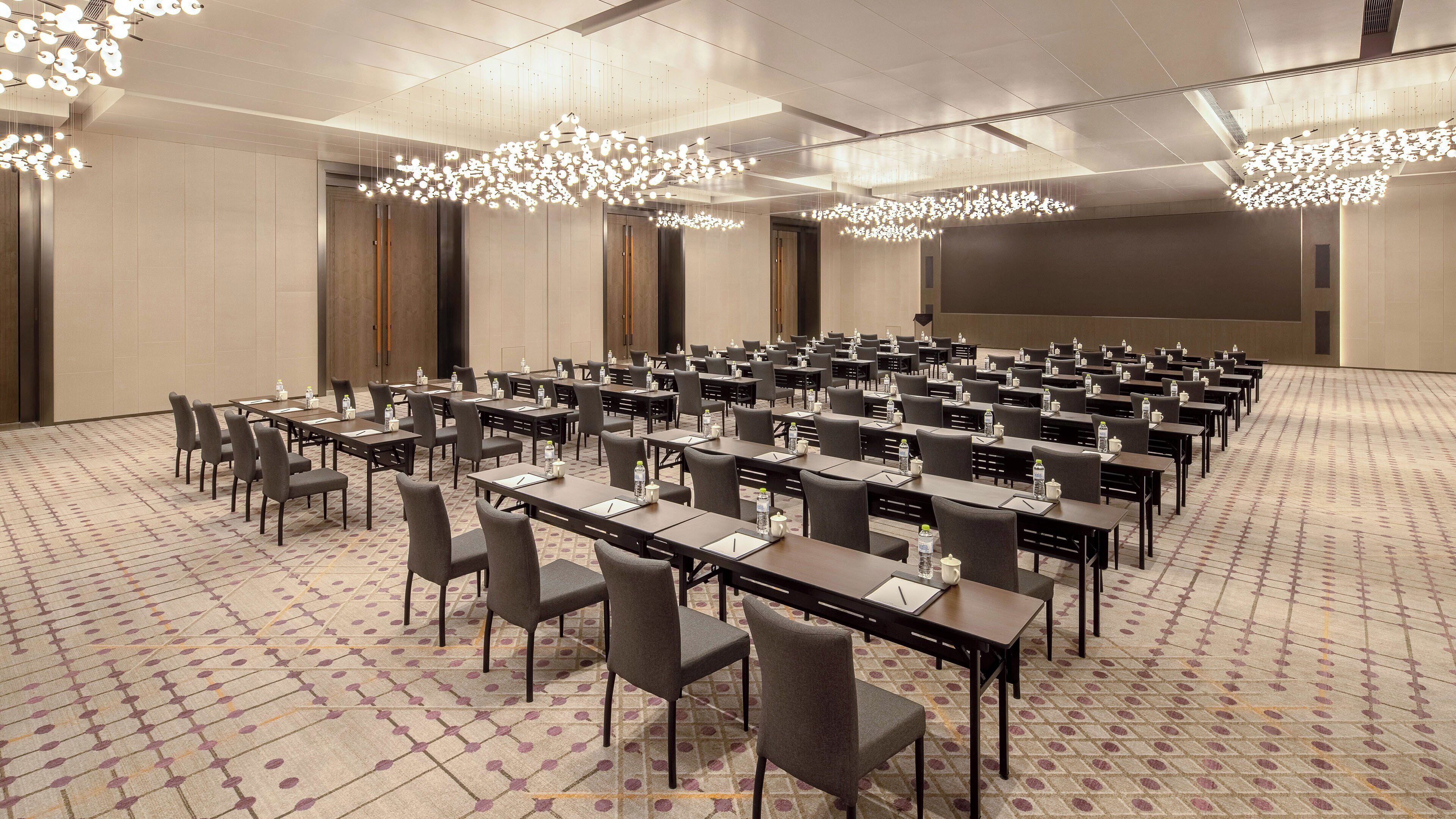 Une salle de conférence avec des rangées de tables et de chaises, équipée de bouteilles d’eau et de blocs-notes, sous de modernes lustres.