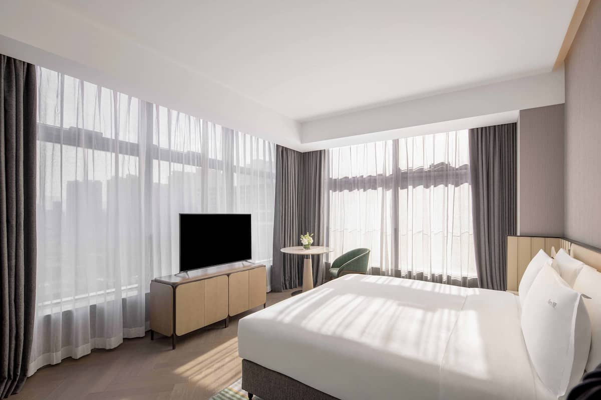 A modern hotel room with a large bed, a flat-screen TV, a small round table with a vase, and a green chair.