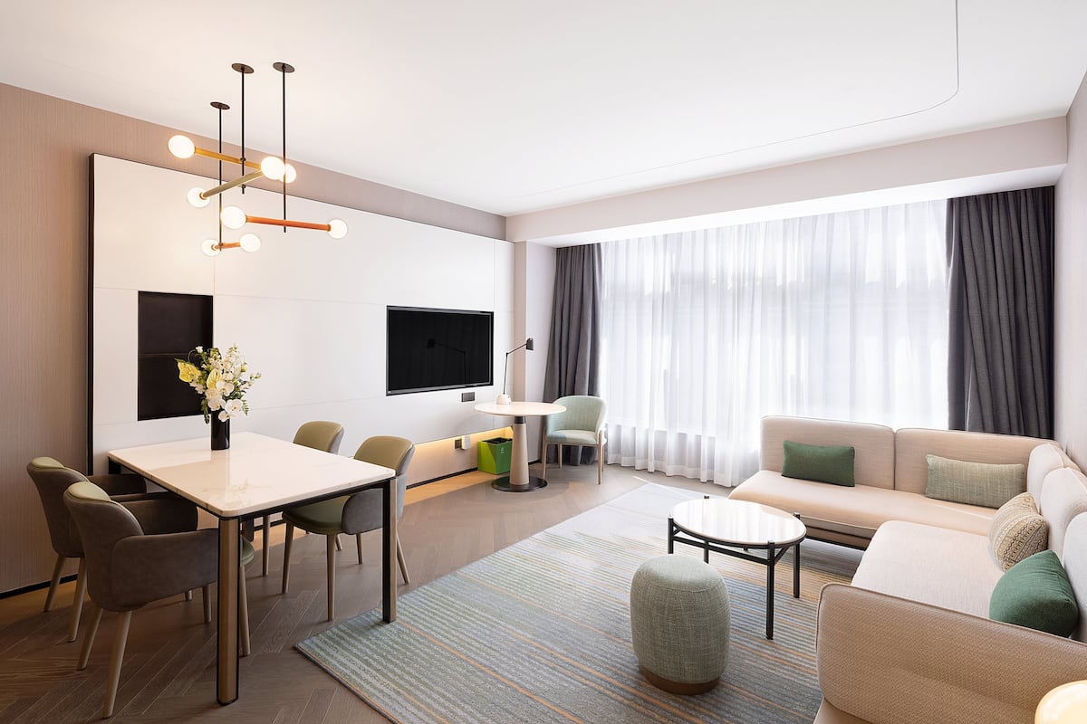A modern hotel room with a dining area, sofa, TV, and large windows.