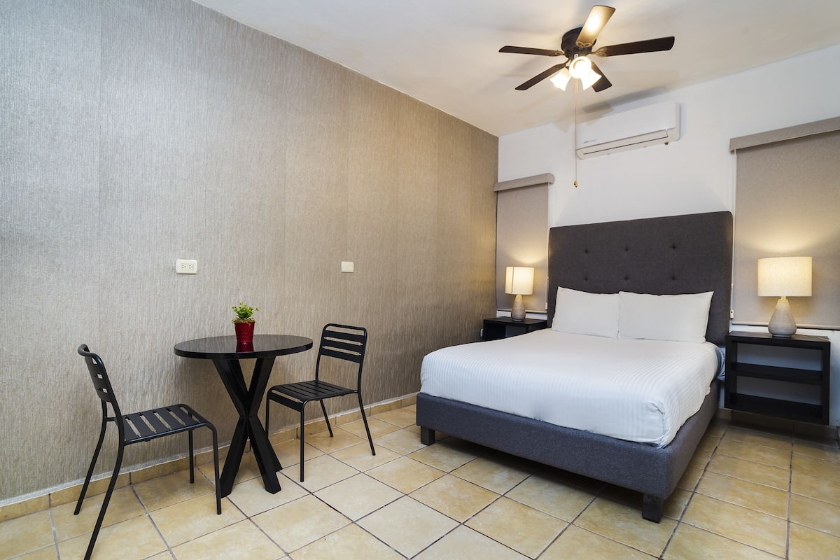 Une chambre d’hôtel comprenant un lit, deux chaises, une petite table, un ventilateur de plafond et deux lampes de chevet.