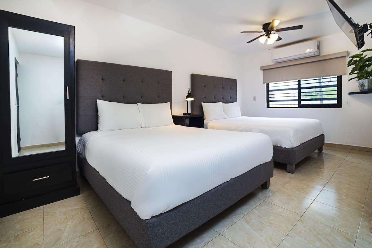 Une chambre avec deux lits, un ventilateur de plafond, un appareil de climatisation, une fenêtre avec des stores et une plante en pot.