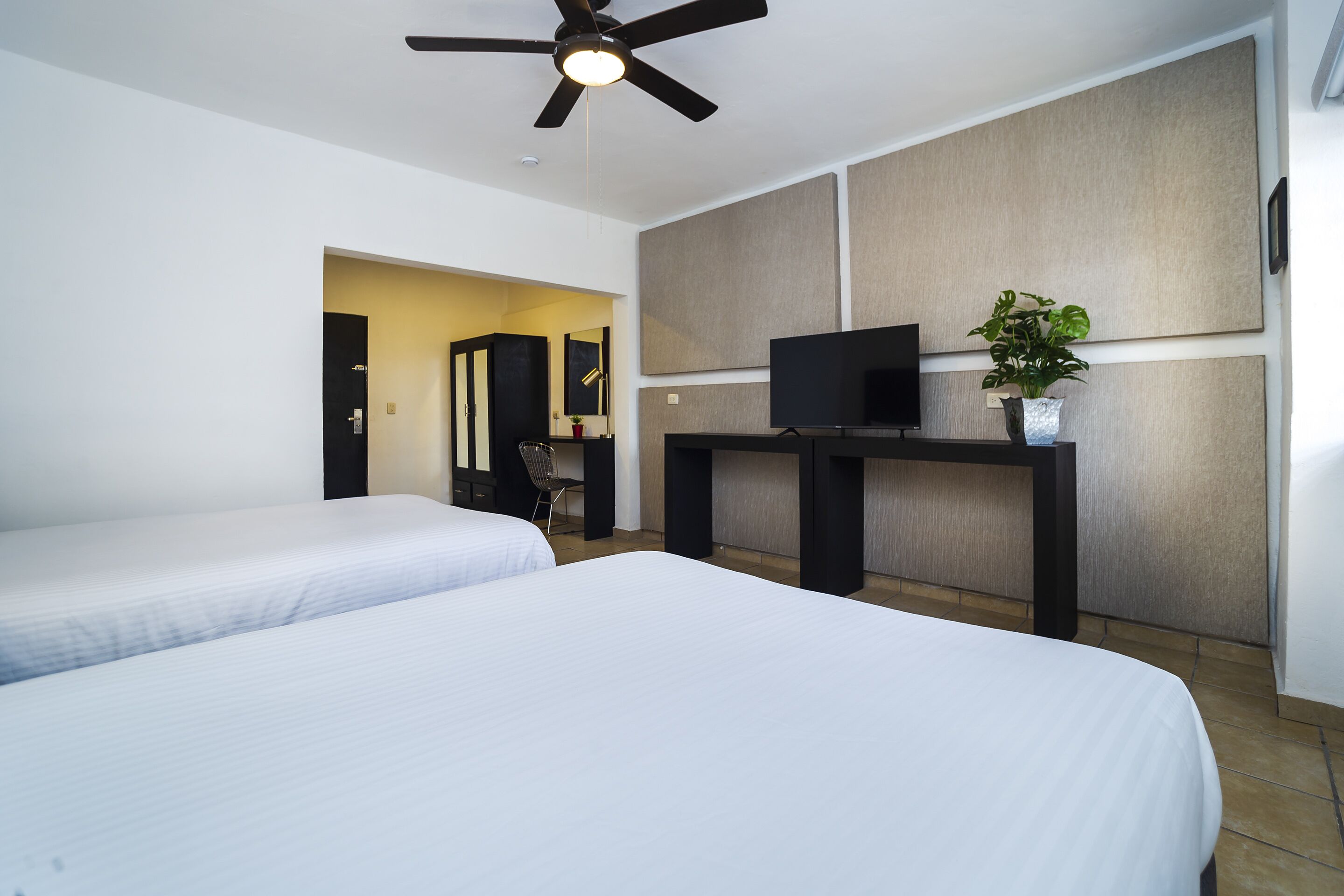 Quarto de hotel com duas camas, uma escrivaninha com televisão, um ventilador de teto e uma planta.