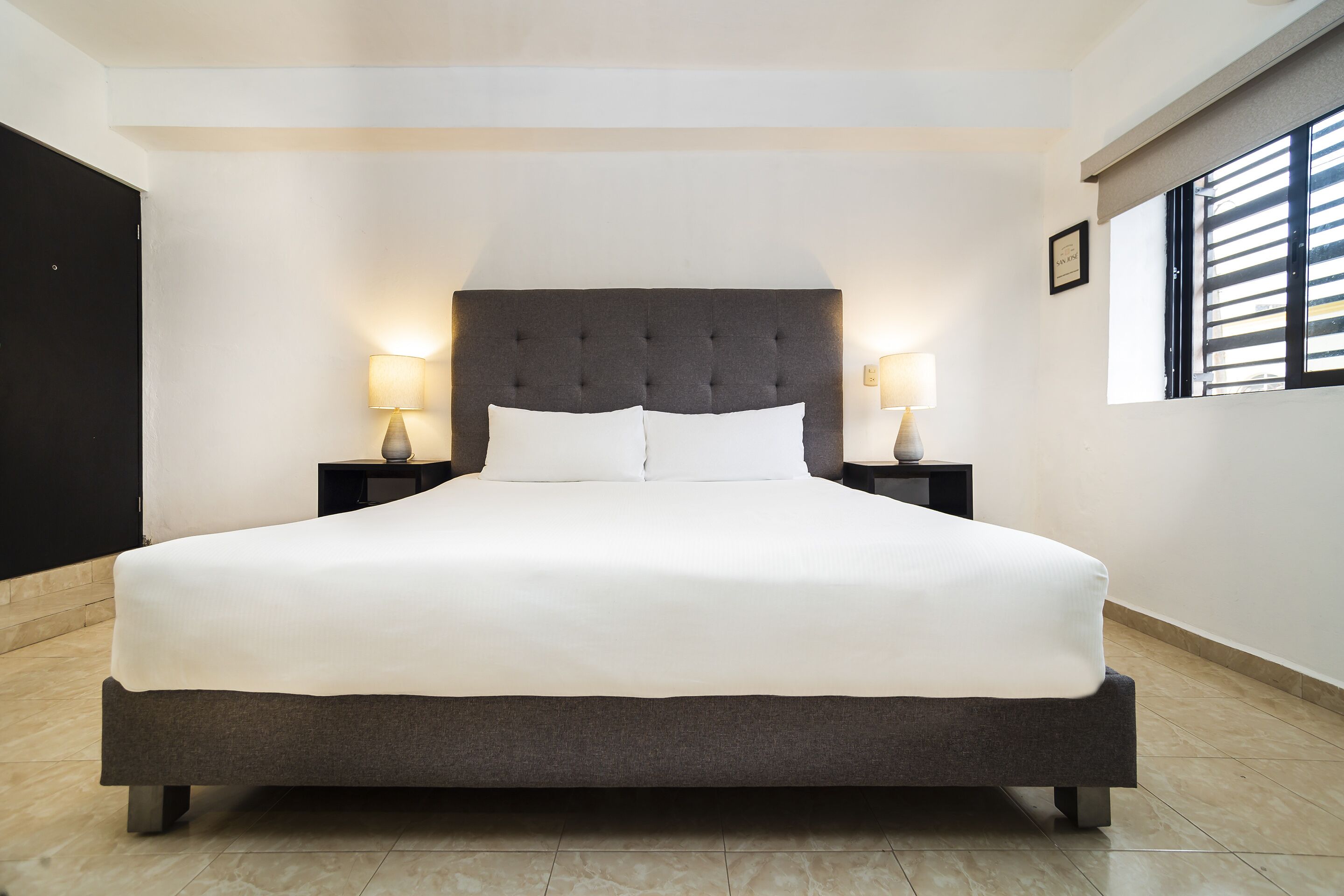 A large bed with a dark headboard and white bedding, flanked by two bedside tables with lamps, in a room with a tiled floor and a window with blinds.
