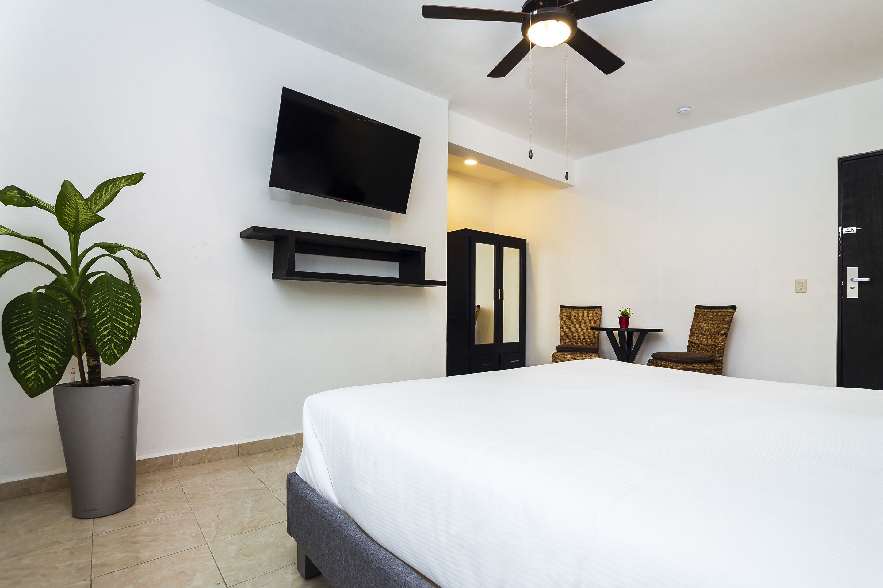 A modern hotel room with a large bed, a flat-screen TV mounted on the wall, a ceiling fan, a potted plant, a small table with chairs, and a wardrobe.