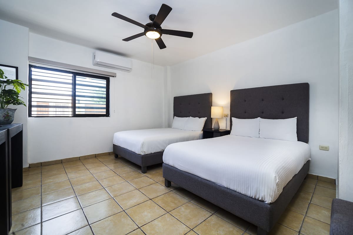 Une chambre d’hôtel avec deux lits, un ventilateur de plafond, un climatiseur, une fenêtre avec des stores et une plante en pot.
