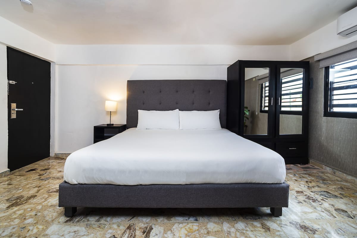 Une chambre d’hôtel moderne avec un grand lit, une table de chevet, une lampe et une fenêtre avec des stores.