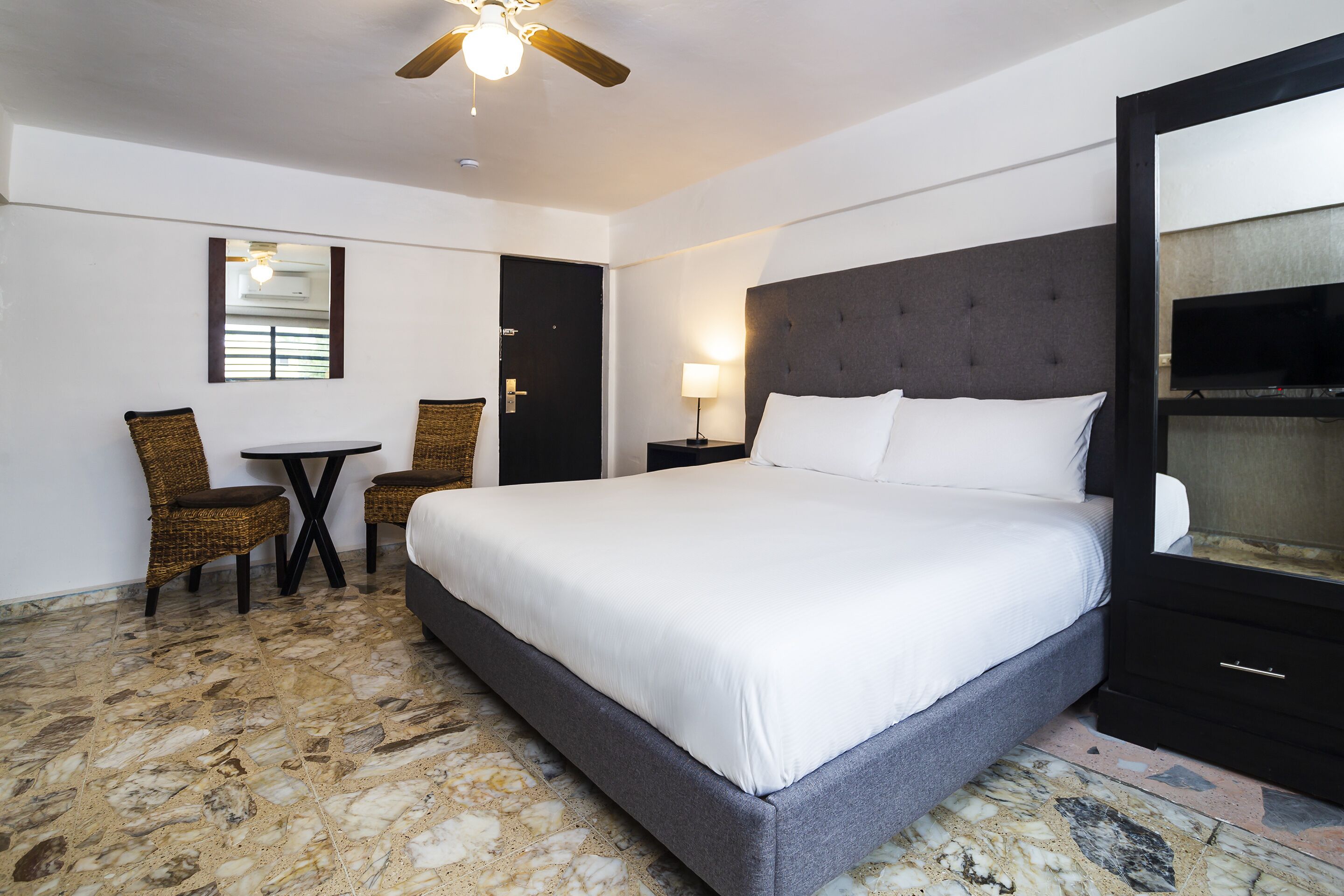 Habitación de hotel con una cama grande, una mesita, dos sillas, un televisor y un ventilador de techo.