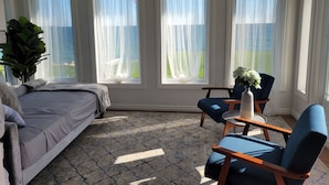 A bedroom with a bed, a chair, a table, and a potted plant. There are three windows with sheer curtains.