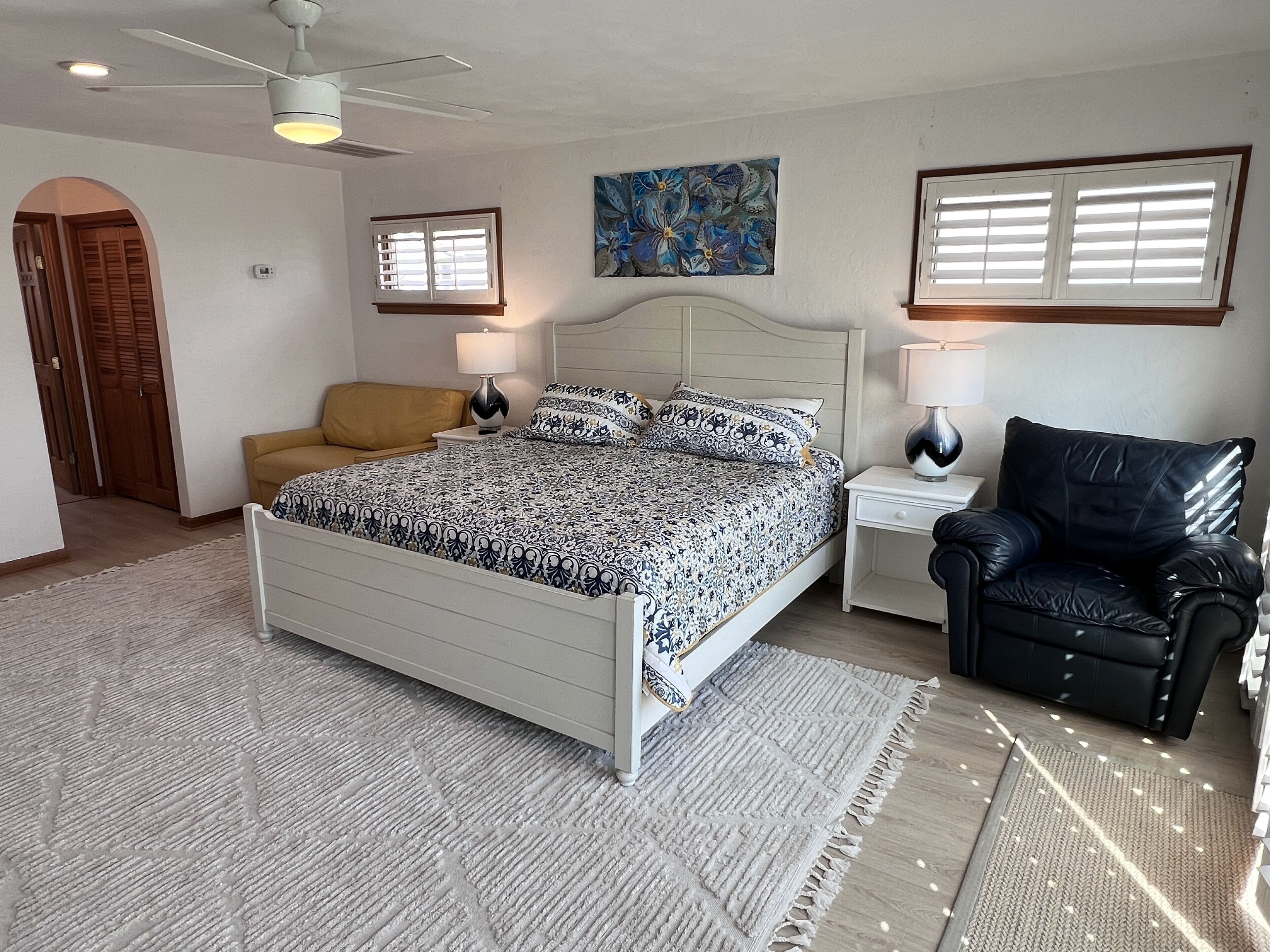 A bedroom with a bed, two bedside tables, a chair, a ceiling fan, and a painting on the wall.