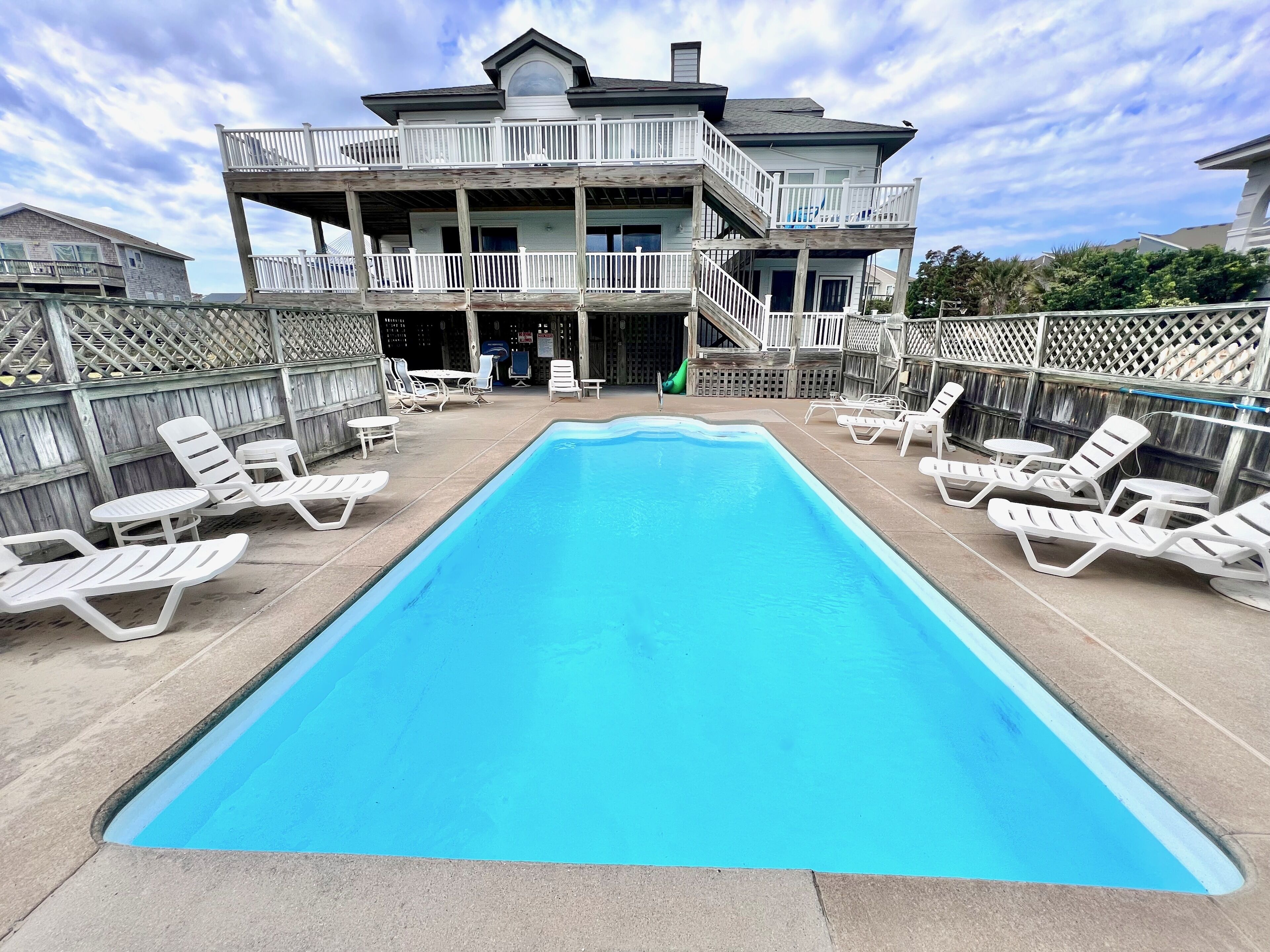 A two-story building with a swimming pool, white deck chairs, and a wooden fence.