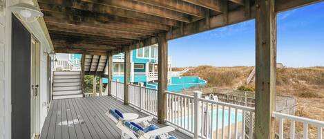 A covered deck with wooden railings, lounge chairs, and a view of a beach and ocean.