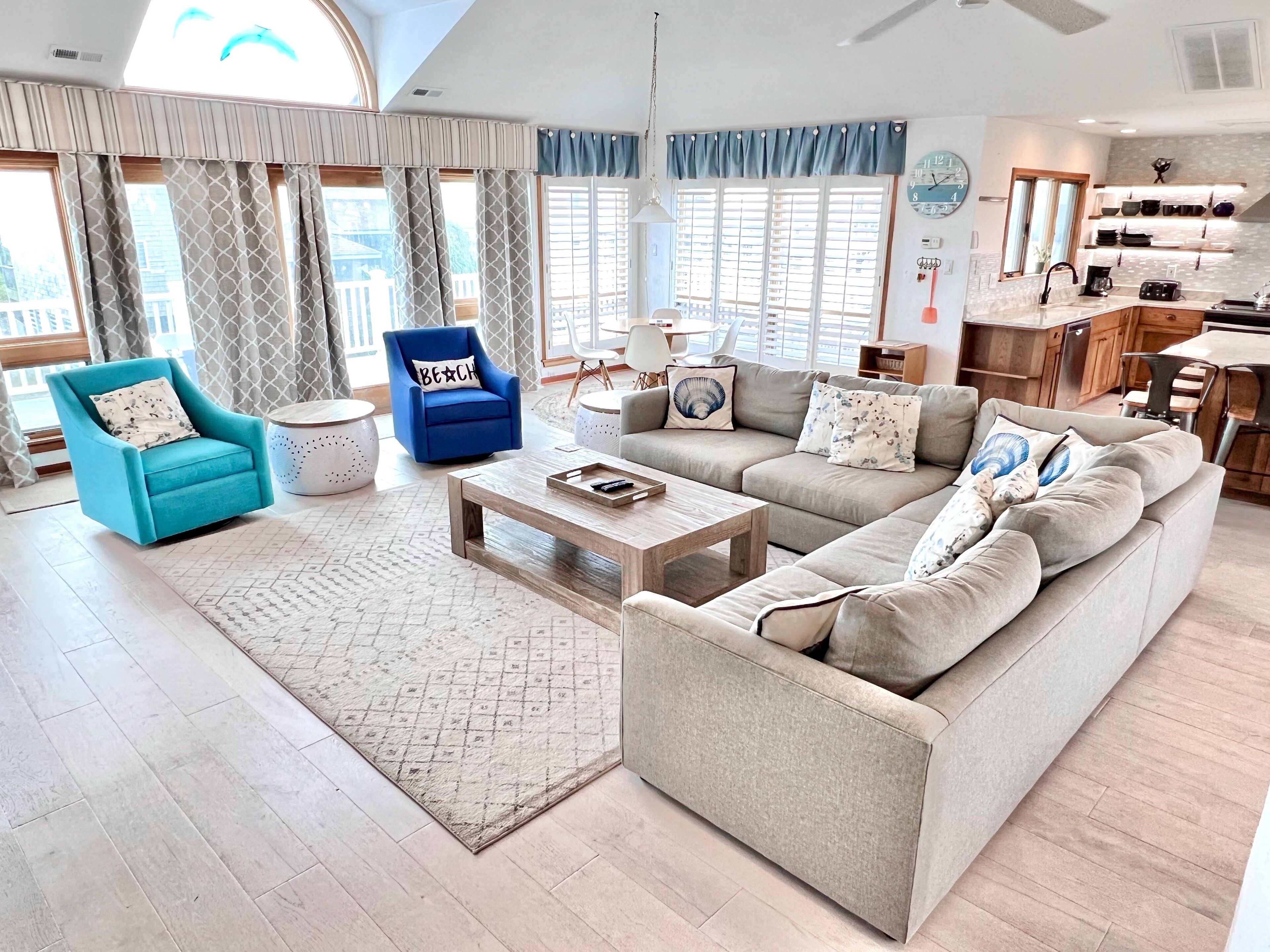 A spacious living room with a large sofa, a blue armchair, and a coffee table. There is a kitchen area visible in the background.