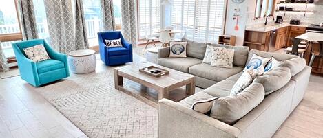 A spacious living room with a large sofa, a blue armchair, and a coffee table. There is a kitchen area visible in the background.
