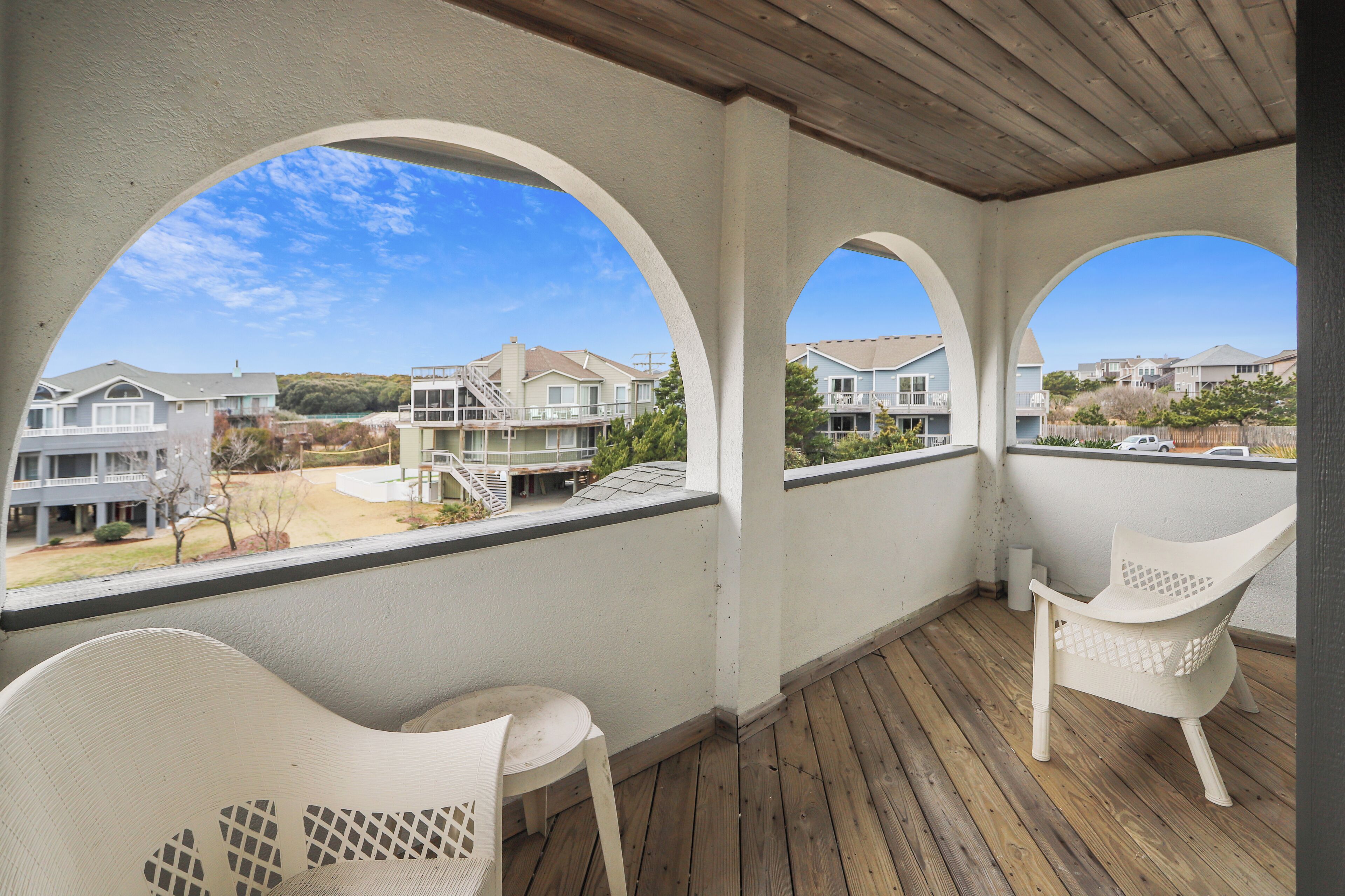 A covered outdoor area with two white chairs and a wooden deck, overlooking a residential complex with multiple buildings and a clear sky.