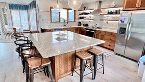 A modern kitchen with a large island, bar stools, wooden cabinets, stainless steel appliances, and pendant lighting. - Ocean Front w/Pool! Hot Tub, Quiet Neighborhood, D0130 (Duck)