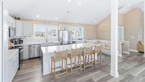 A modern kitchen with white cabinetry, a central island with bar stools, and a view of the beach. - Modern, Community Pool, Quick walk to beach! Hot tub! NH4912 (Nags Head)