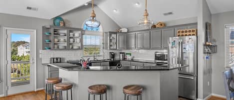 Una cocina moderna con gabinetes grises, una isla central con taburetes, electrodomésticos de acero inoxidable e iluminación colgante.