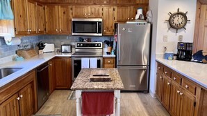 A modern kitchen with wooden cabinets, stainless steel appliances, a granite countertop, and a clock on the wall. - Steps to the Sand! Ocean Views! Hot tub! Specials! Dogs! KD0223 (Kill Devil Hills)