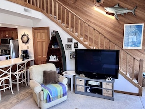 Una sala acogedora con un televisor de pantalla plana grande, un sillón cómodo, una escalera de madera y una zona de cocina al fondo.